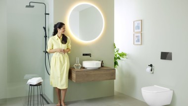 Ženska v rumeni obleki stoji pred metino zeleno kopalnico s pohištvom in kopalniško keramiko Geberit ter črnimi pipami