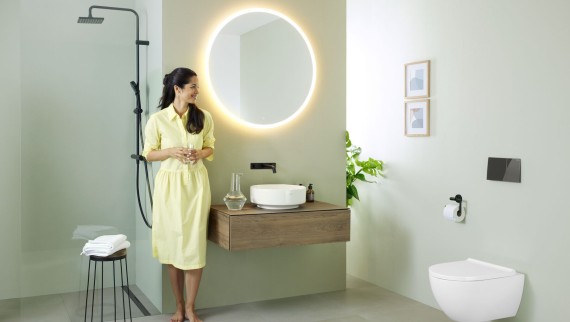 Ženska v rumeni obleki stoji pred metino zeleno kopalnico s pohištvom in kopalniško keramiko Geberit ter črnimi pipami