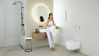 Ženska se naslanja na umivalnik v kopalnici s straniščem s prho Geberit AquaClean Sela in umivalnikom Geberit VariForm ter pohištvom