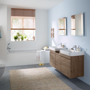 Družinska kopalnica s svetlo modro steno in kopalniškim pohištvom v barvi hikorja, omarico z ogledalom, aktivirno tipko in keramiko Geberit